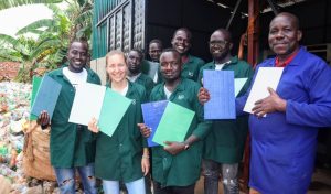 A group of eight individuals, wearing green and blue work jackets, proudly display sheets of recycled material. They are standing outside a recycling facility, with bags of sorted plastic bottles visible in the background. The diverse group, consisting of both men and women, smiles at the camera.