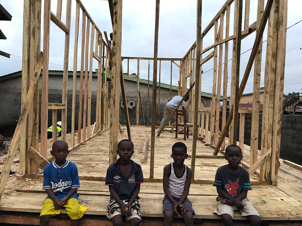 Four young boys sit on a wooden platform that is part of the framework for a new building under construction. The structure is made of wooden beams, forming the skeleton of walls and a floor. The boys, who appear to be around 5-7 years old, are dressed in casual clothes and look directly at the camera.