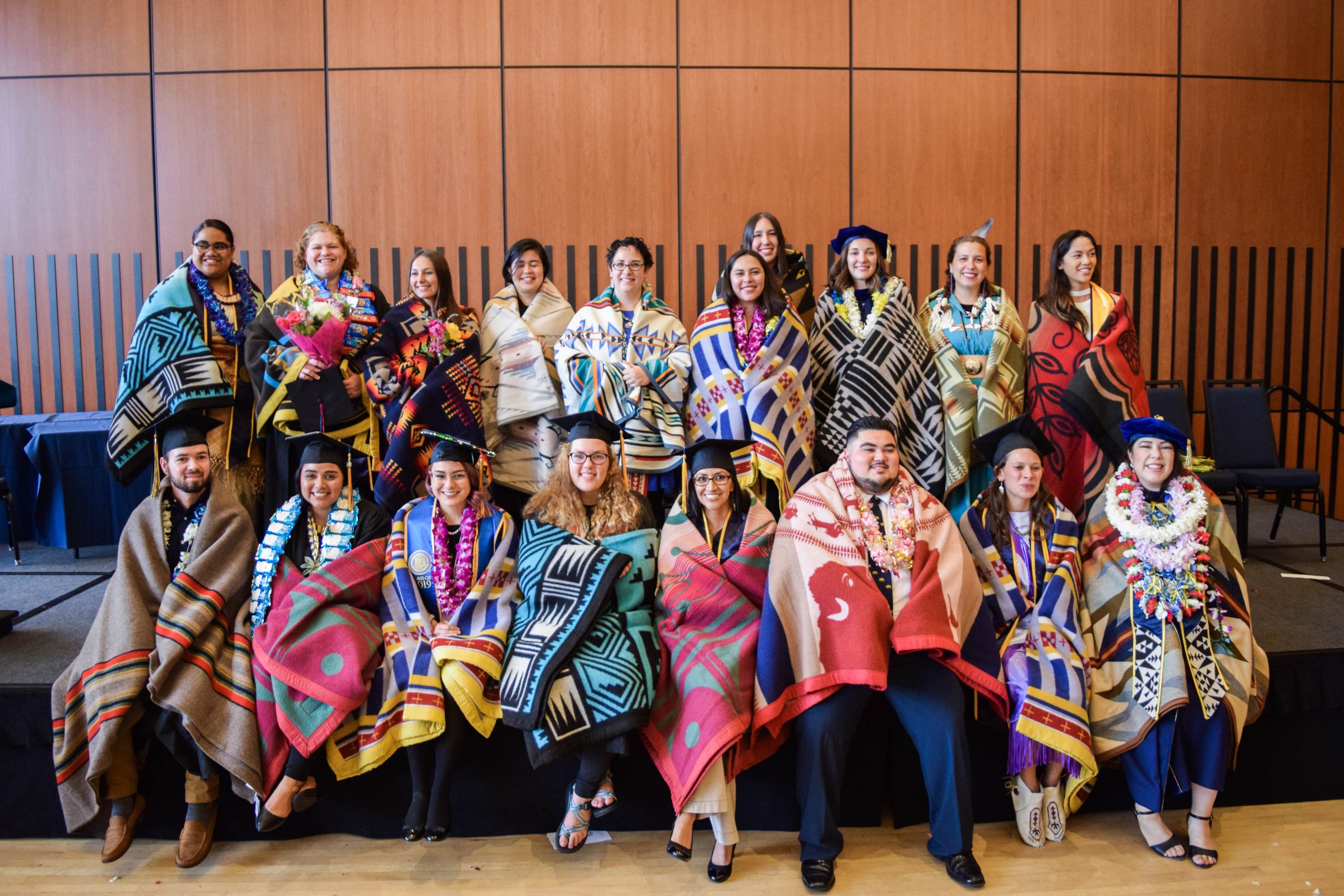 A group of graduates pose for a group photo in a large hall. They are wearing graduation caps and are wrapped in colorful, patterned blankets