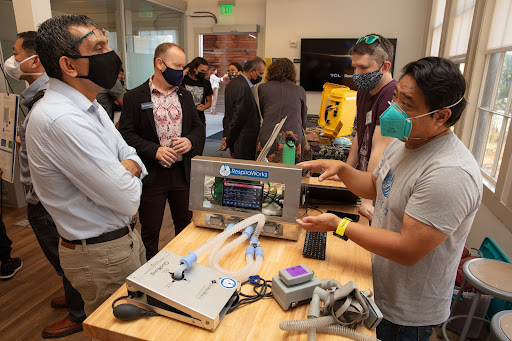Several people, all wearing face masks, are gathered around a table in a bright, well-lit room with large windows. On the table, there is medical equipment, including a device with tubes and a screen displaying "RespiraWorks."