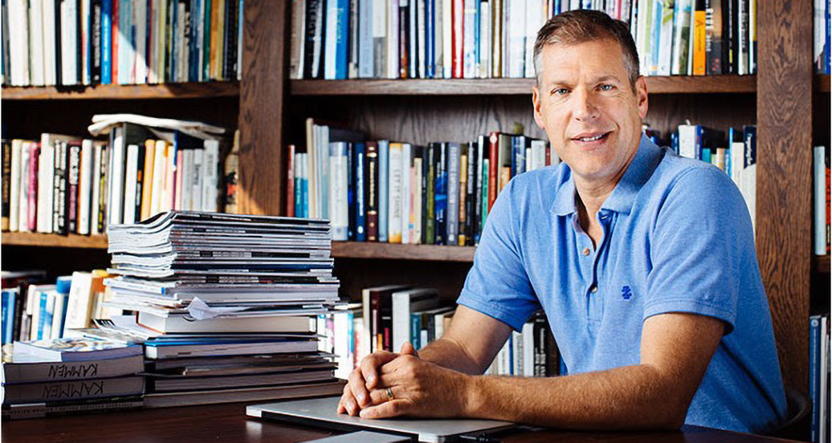 A man in a blue polo shirt is seated at a desk in front of a large bookshelf filled with books. He is smiling and has his hands resting on the desk, which is cluttered with stacks of books and magazines.