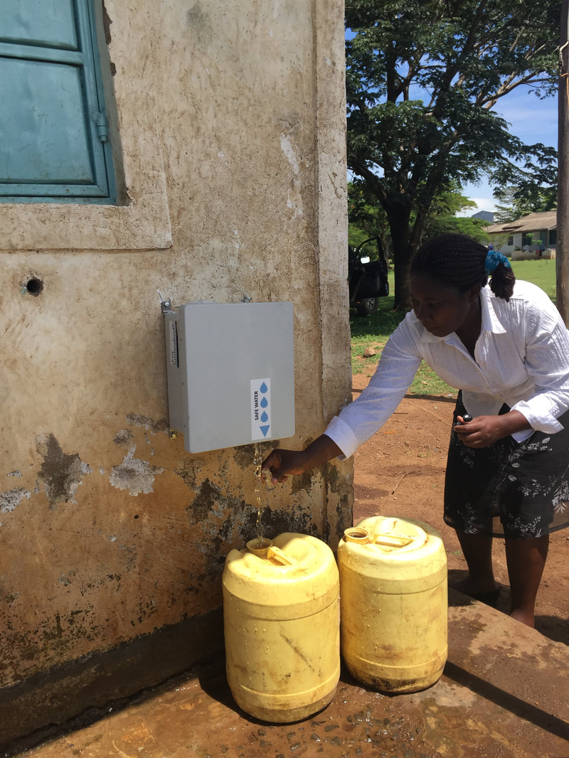 A woman is filling yellow water containers from a water dispensing unit attached to the wall of a building. The building has a blue window and a worn-out exterior. The woman is wearing a white shirt and a patterned skirt.