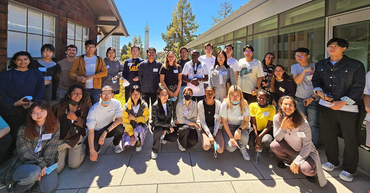 A group of diverse students and faculty pose together outdoors on a sunny day. They are standing on a patio with a mix of smiles and casual postures, wearing name tags and a variety of casual outfits.