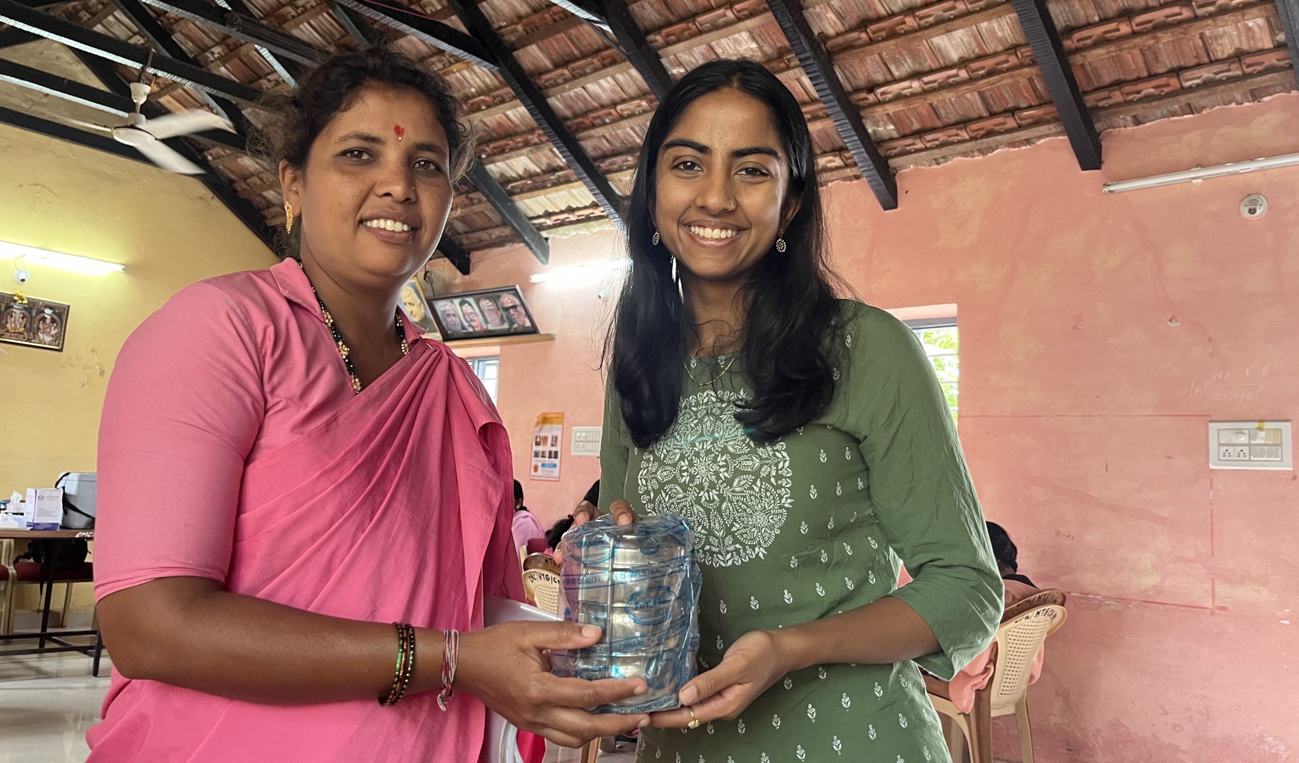 A woman in a pink sari and a woman in a green dress are standing together, smiling at the camera. The woman in the green dress is holding a wrapped item.