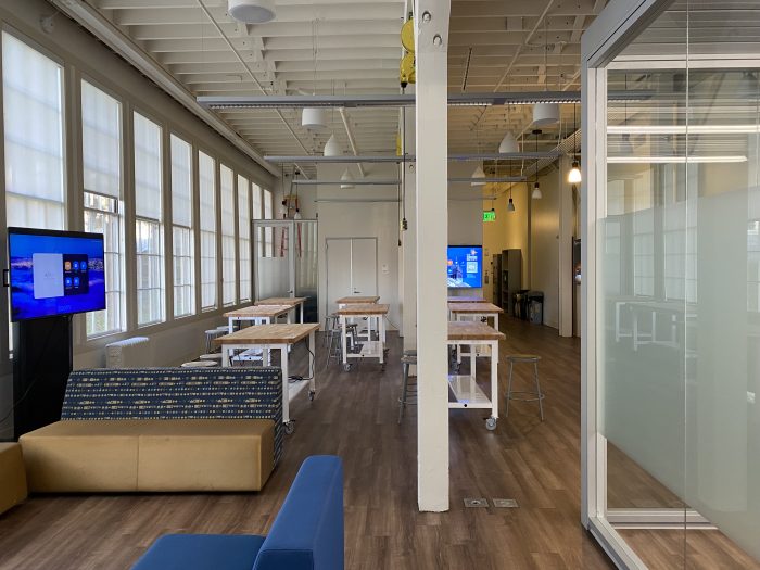 An open and versatile workspace filled with natural light from the large windows along one side of the room. The space features several large wooden tables on white metal frames with wheels, paired with gray metal stools. On the left side, there are comfortable seating options, including a blue and yellow patterned couch. Large screens are positioned for presentations or virtual meetings.