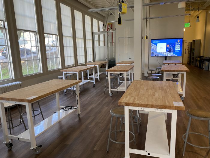 CoLab - Main: A well-lit workspace featuring several large wooden tables on white metal frames with wheels. Each table is paired with gray metal stools, providing seating for multiple users. The room has large windows that allow ample natural light. In the background, a large screen displays content related to Berkeley.