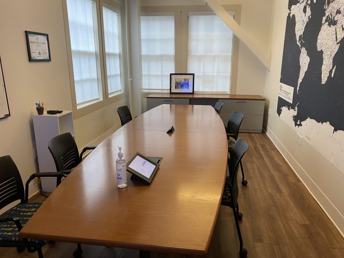 CoLab - B120B: A modern conference room with a long wooden table surrounded by black office chairs. The room features large windows with frosted glass, allowing natural light to fill the space. On the table, there is a bottle of hand sanitizer and a tablet device. The walls are adorned with a large world map and a framed certificate.