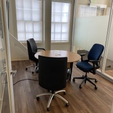 CoLab - B120C: A small meeting room with wooden floors, featuring a round wooden table and three office chairs. The room has large windows with frosted glass providing natural light.