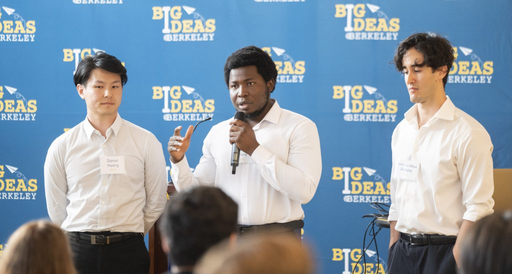 Three men are presenting at a "Big Ideas @ Berkeley" event. The man in the center is speaking into a microphone, while the other two stand attentively beside him. They are dressed in white shirts and dark trousers, and each has a name tag. The backdrop displays the event's logo repeatedly.