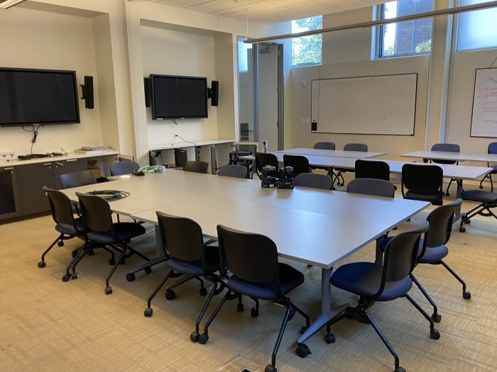 Room B100 A/B: A well-equipped, modern conference room featuring multiple tables arranged for group collaboration. The room includes several flat-screen monitors mounted on the walls, along with whiteboards for presentations and brainstorming sessions. Chairs on wheels are positioned around the tables for easy movement. The room is brightly lit with natural light coming through the windows.