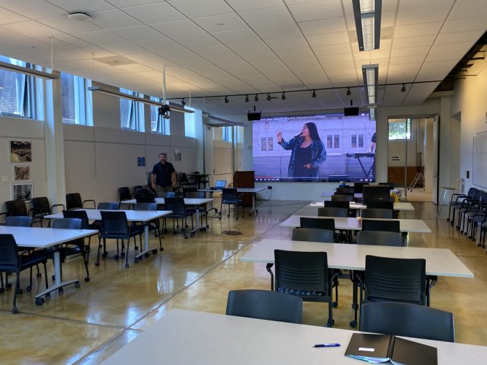 A spacious, well-lit conference room or classroom equipped for presentations and collaborative work. The room features multiple tables and chairs on wheels, arranged to facilitate group interactions. A large projection screen at the front displays a video of a speaker, suggesting the room is set up for a presentation or lecture. The walls have high windows that allow natural light to flood the space, creating an open and inviting atmosphere. A person stands near the back of the room, emphasizing the room's size and flexibility for various configurations. The polished concrete floor adds to the modern and clean aesthetic of the room.
