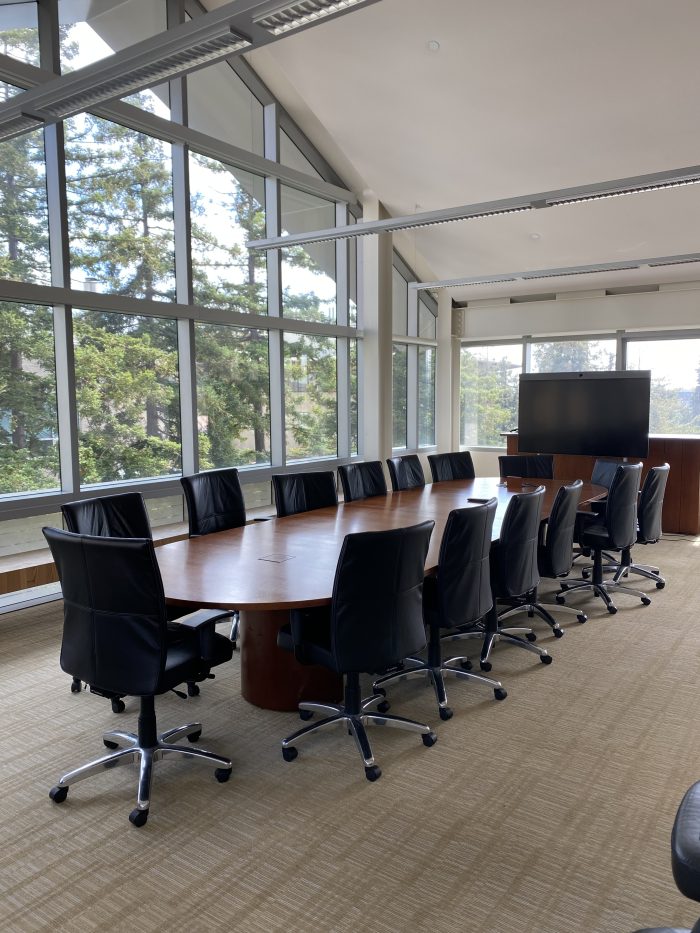 A modern conference room with a large wooden table surrounded by black leather chairs on wheels. The room is bright and airy, with floor-to-ceiling windows offering a view of green trees outside. The high ceiling and ample natural light. At the far end of the room, there is a large screen.